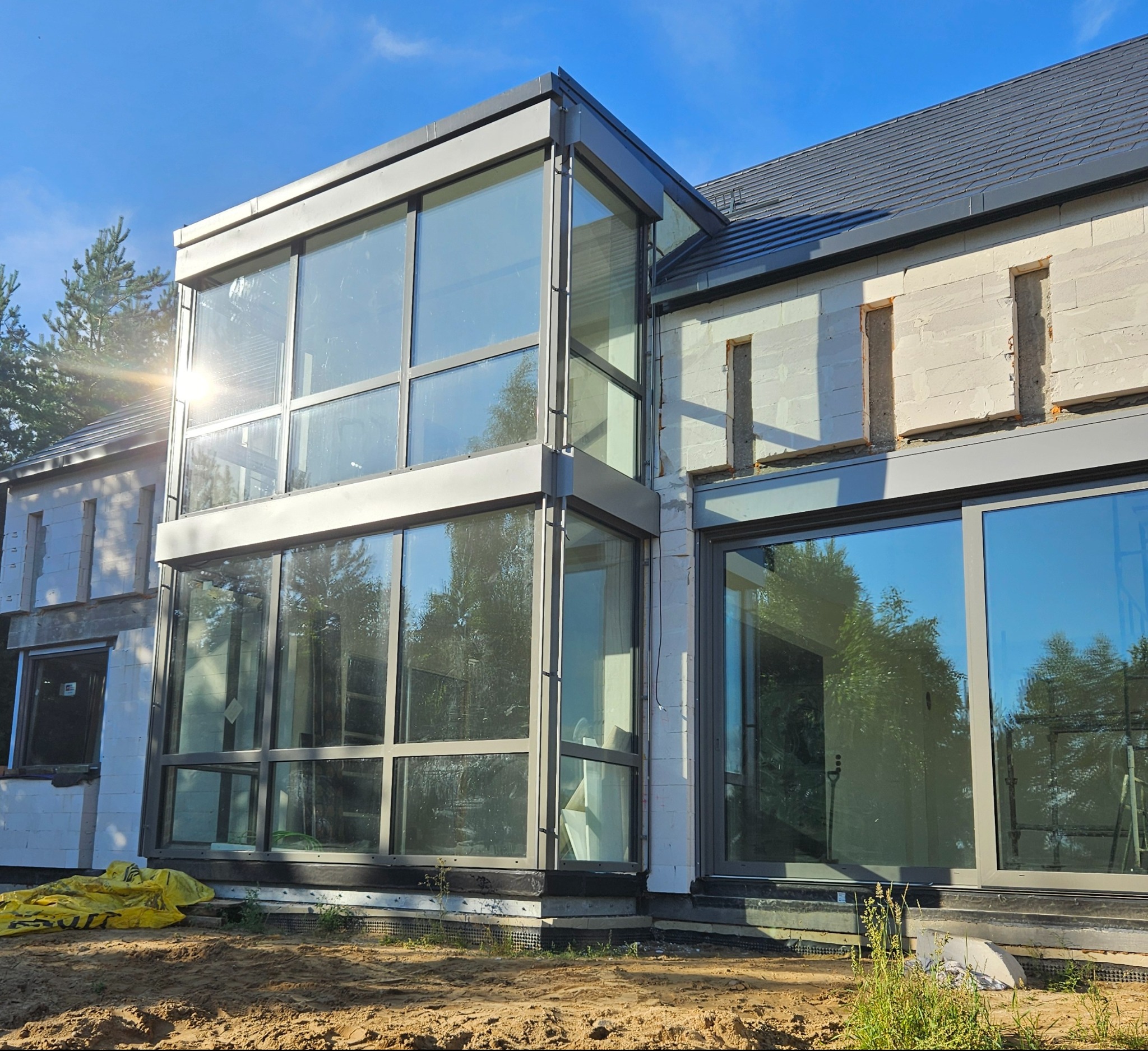 Nowoczesna bryła domu w trakcie budowy z dużymi oknami aluminiowymi, widoczne pustaki i elementy konstrukcyjne, jasne światło słoneczne.