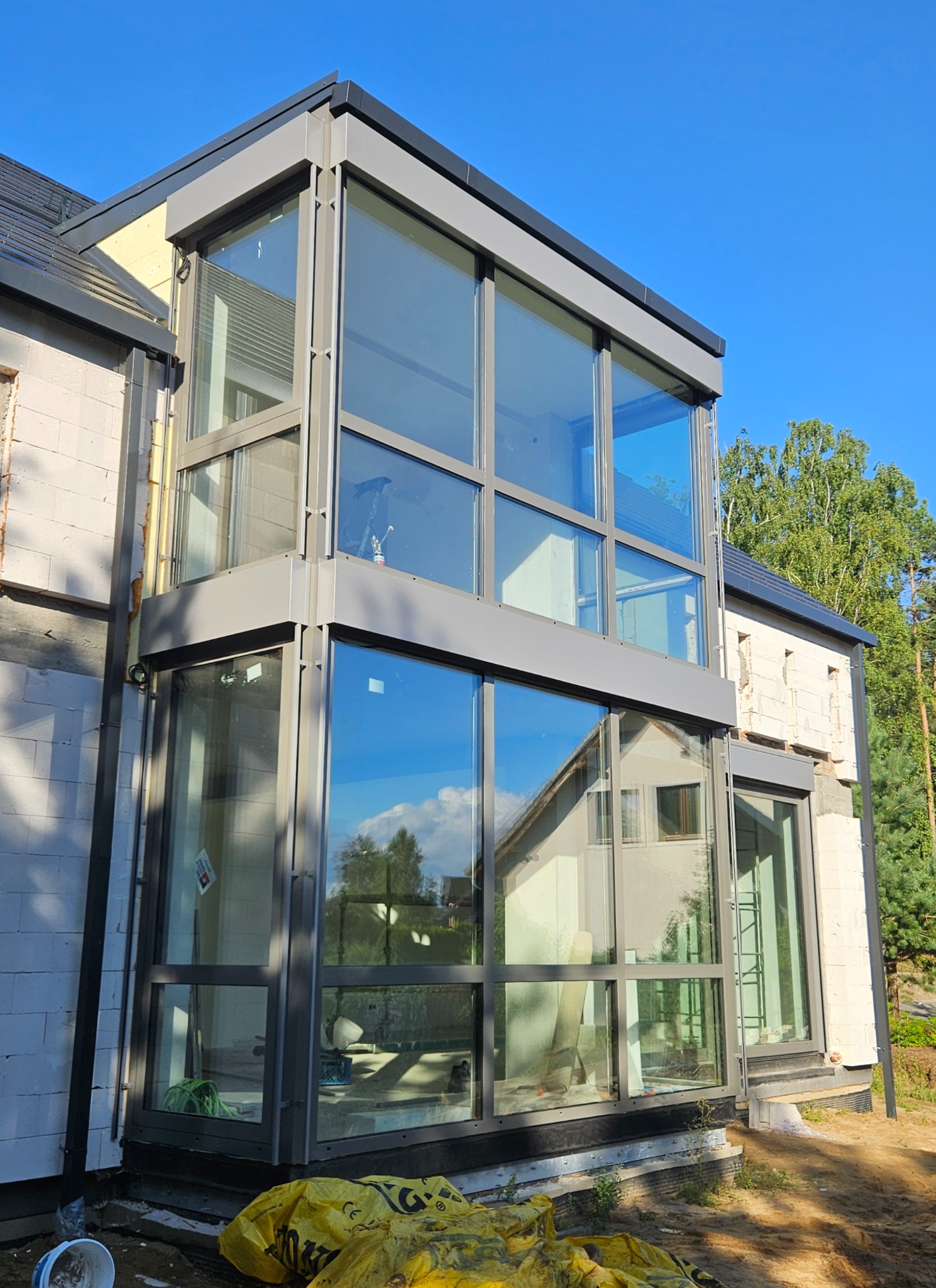 Dwukondygnacyjny, przeszklony wykusz z szarą ramą, dobudowany do budynku z białego bloczka. Widoczne odbicie nieba i drzew w szybach. Na dole żółte worki budowlane.