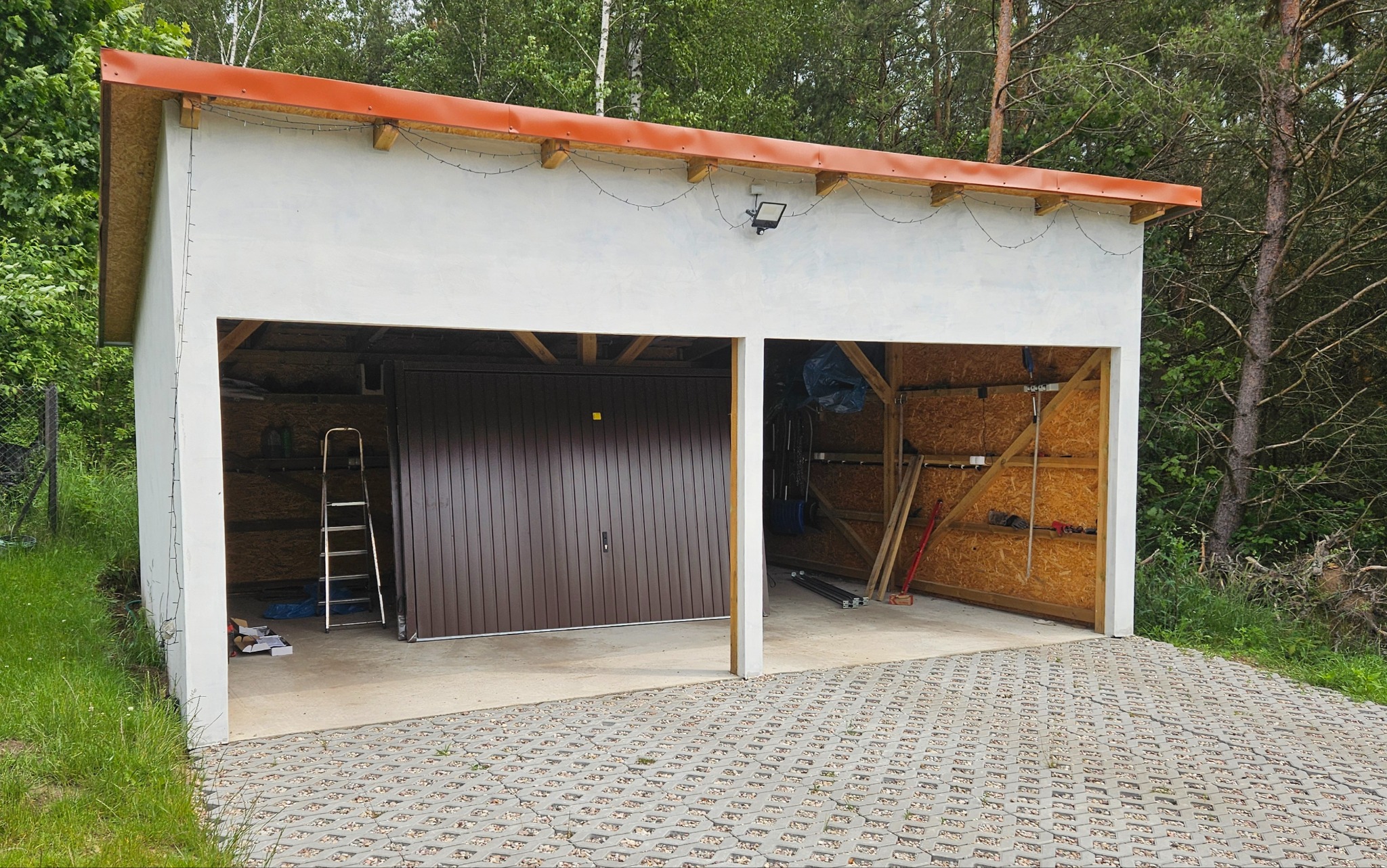 Dwustanowiskowy garaż z brązową bramą segmentową po lewej stronie, oświetleniem zewnętrznym i kostką ażurową przed wjazdem, otoczony zielenią.