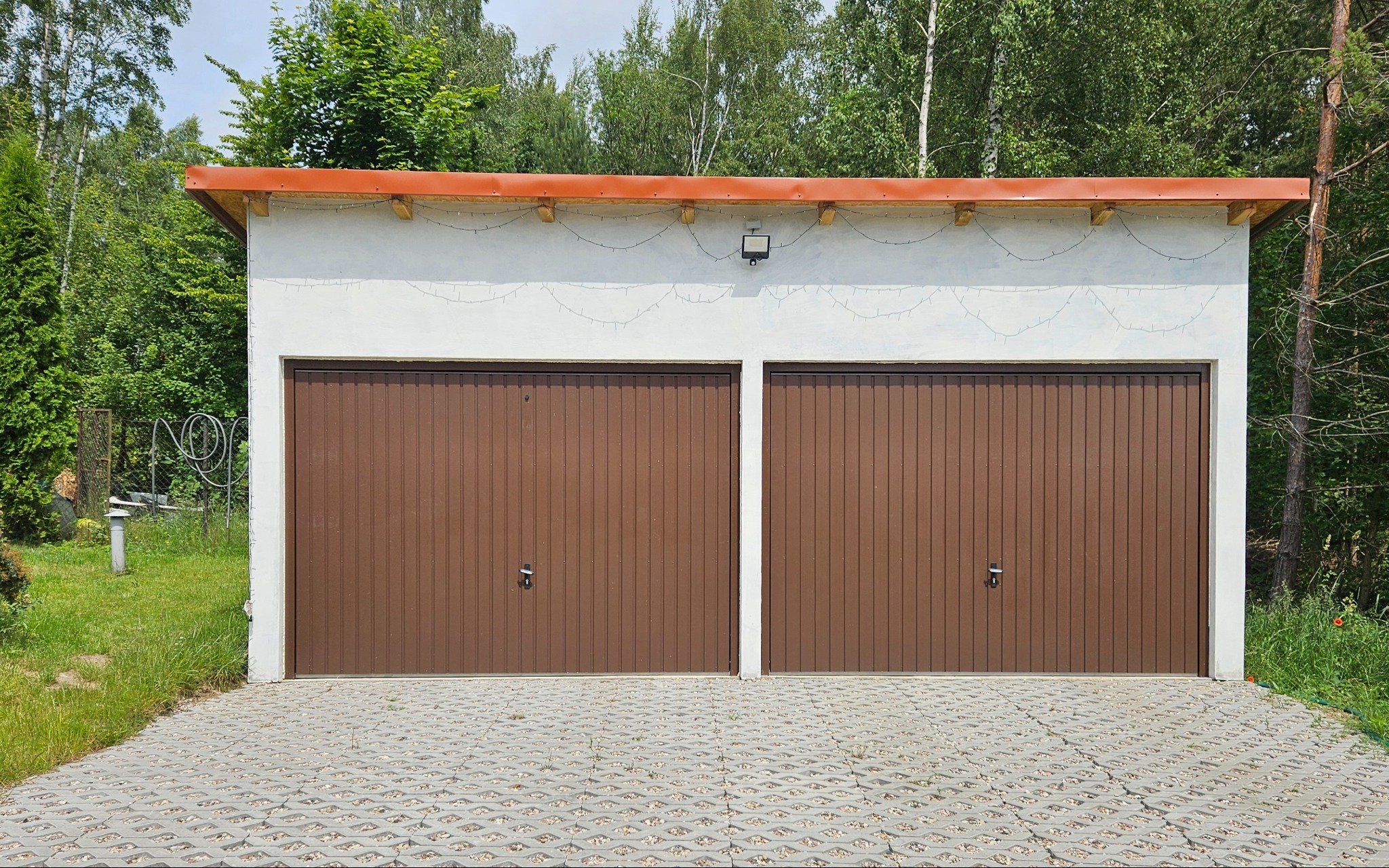 Dwustanowiskowy garaż z brązowymi bramami segmentowymi w Słupsku. Biały budynek z oświetleniem zewnętrznym i dekoracyjnymi lampkami. Kostka ażurowa przed wjazdem.