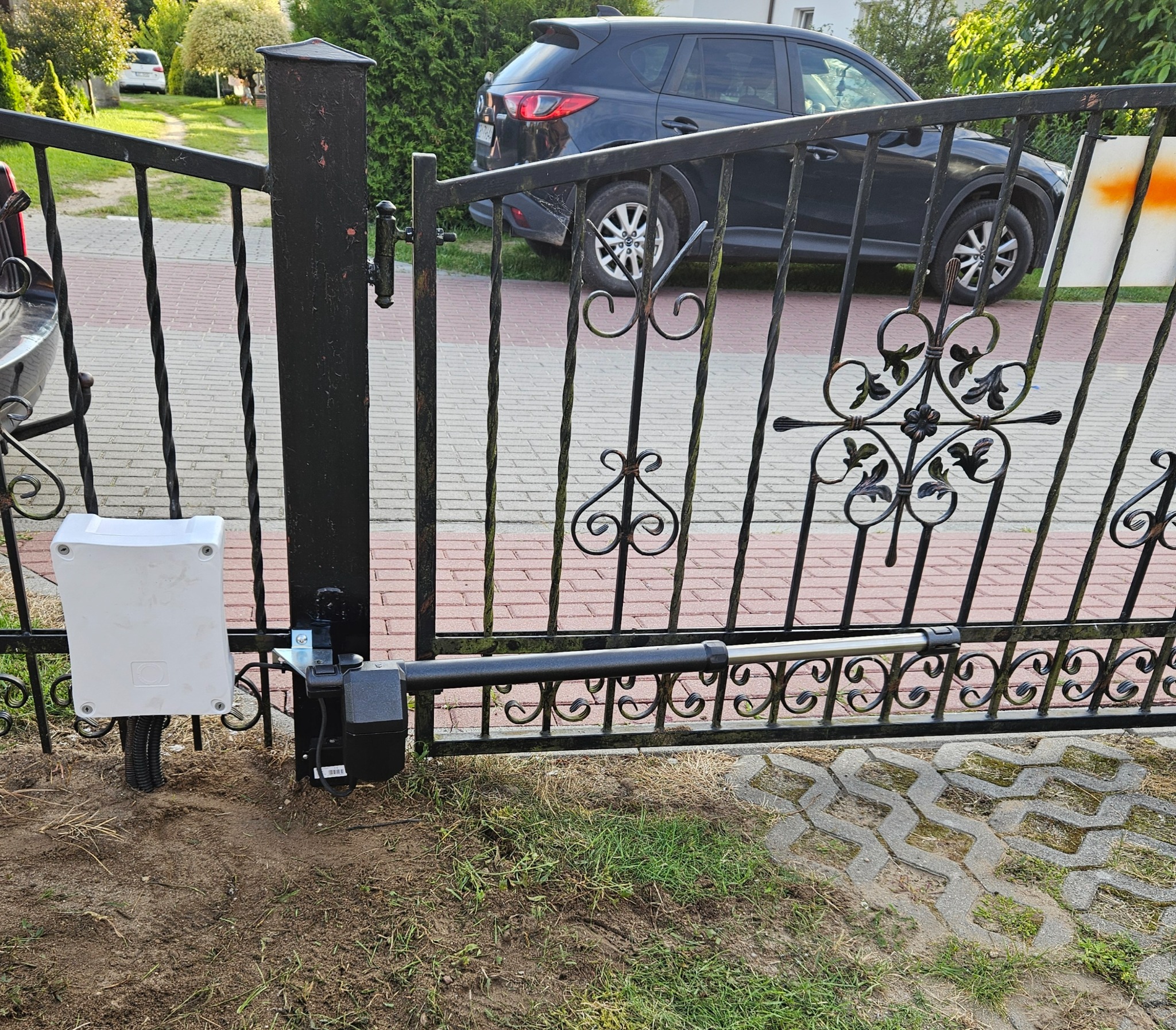 Czarna, metalowa brama z automatyką, widoczny siłownik i skrzynka sterująca. W tle zaparkowany samochód i kostka brukowa.