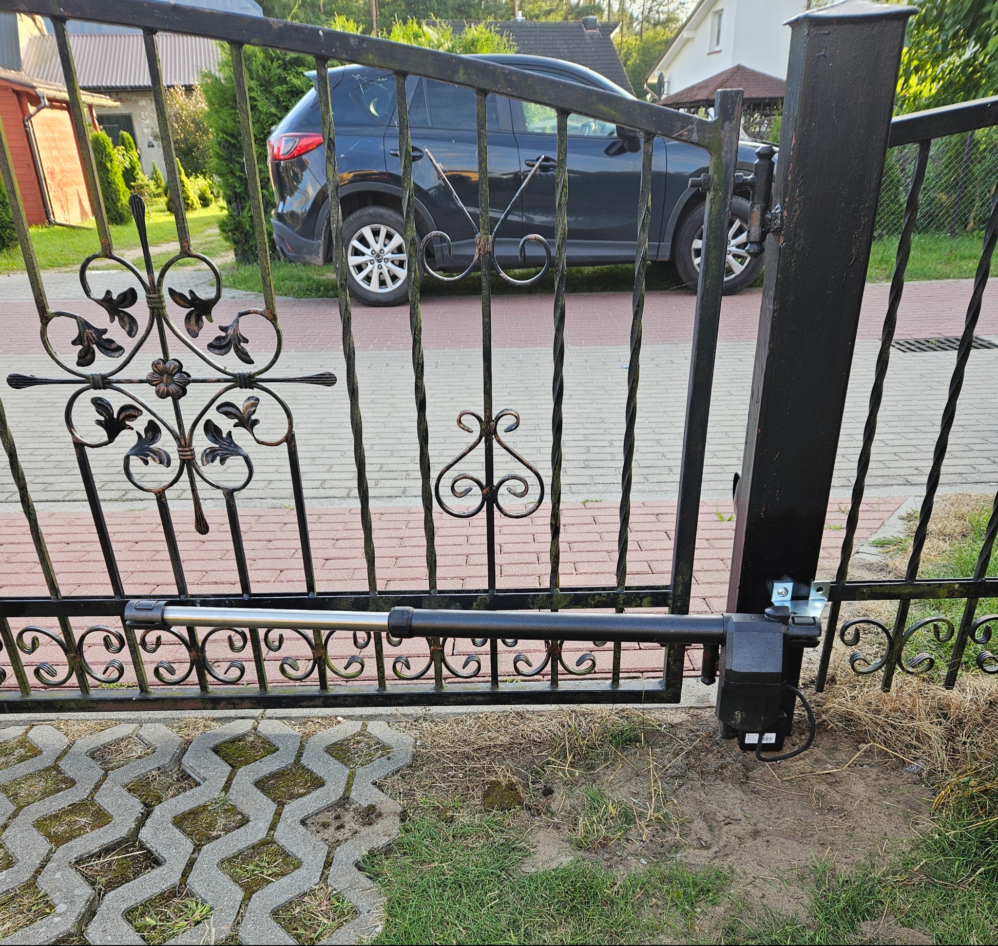 Kuta brama wjazdowa z automatyką, widoczny siłownik. W tle zaparkowany samochód i dom. Kostka brukowa na podjeździe.