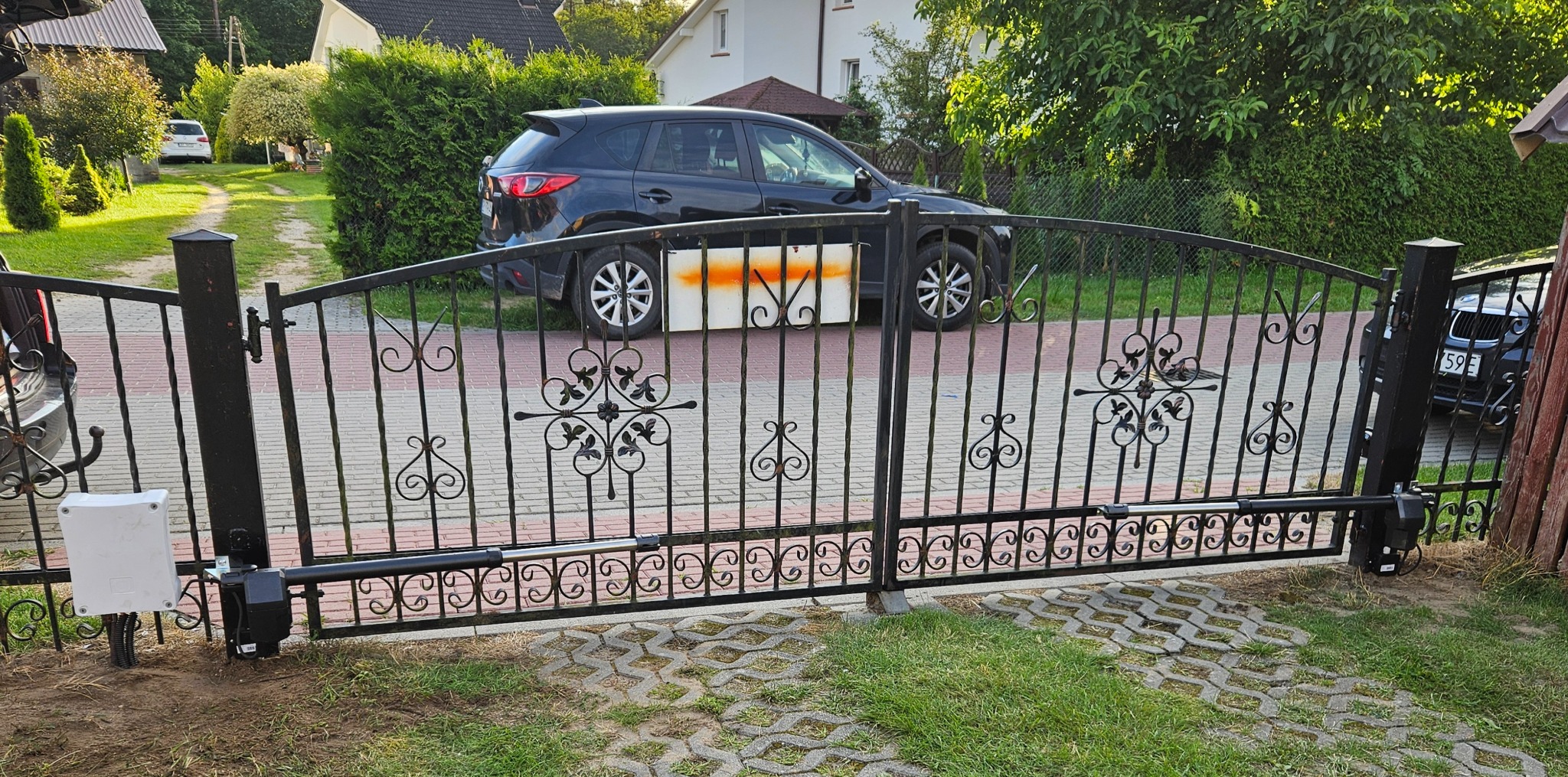 Kuta brama wjazdowa z automatyką, widoczny siłownik i skrzynka sterująca. W tle zaparkowany samochód i dom jednorodzinny.