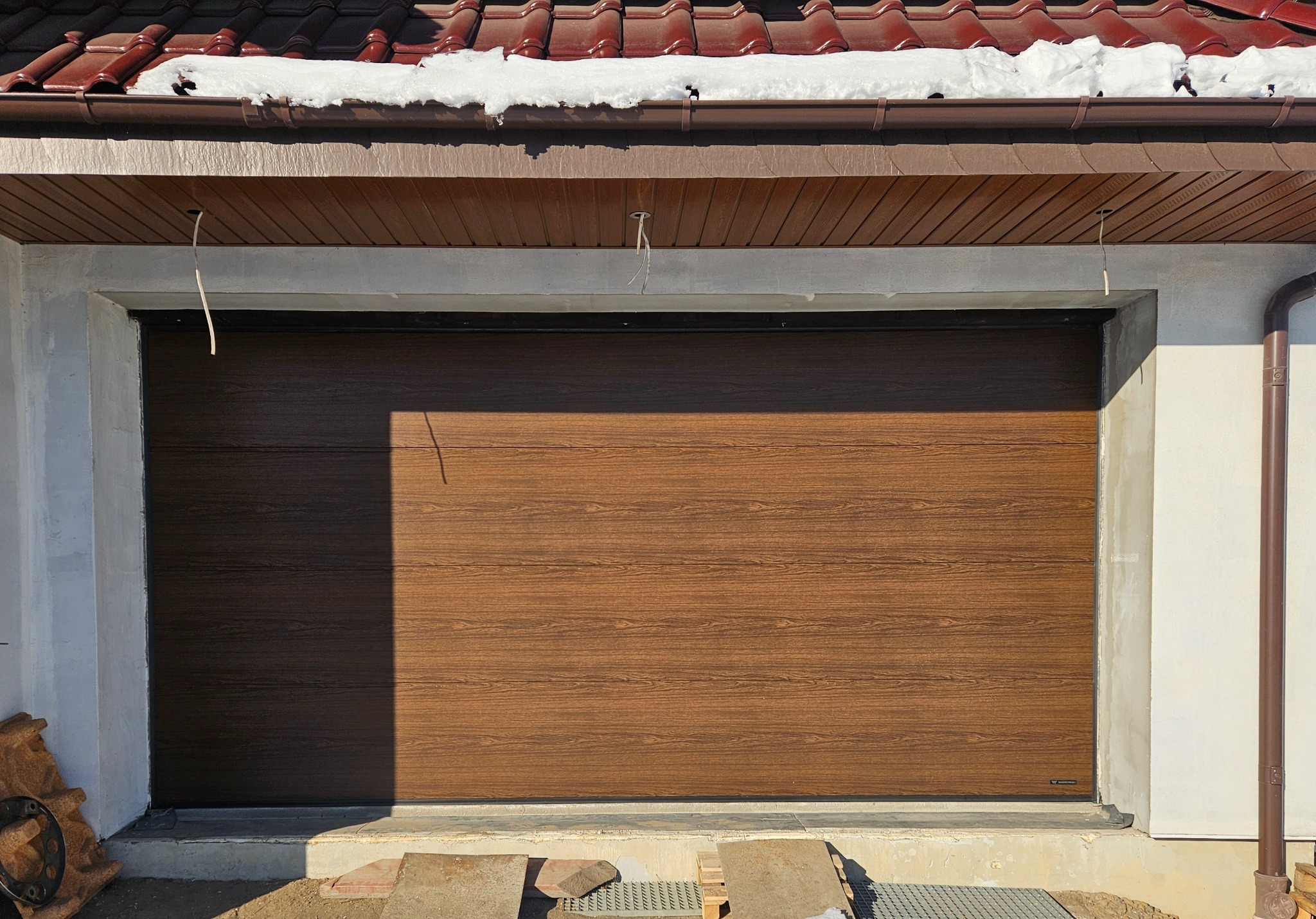 Zamknięta, segmentowa brama garażowa w kolorze drewna, z widocznym kablem wystającym z sufitu. Częściowo ośnieżony dach i rynna. Widoczne płyty chodnikowe.