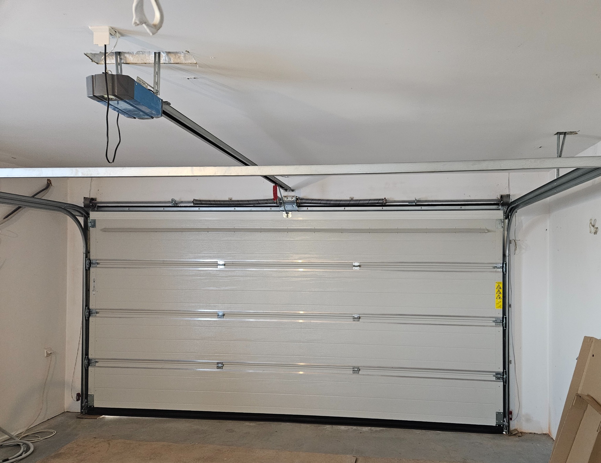 Zamontowana brama garażowa segmentowa z napędem w Słupsku. Widoczne sprężyny i prowadnice. Brama w kolorze jasnym, z poziomymi przetłoczeniami.