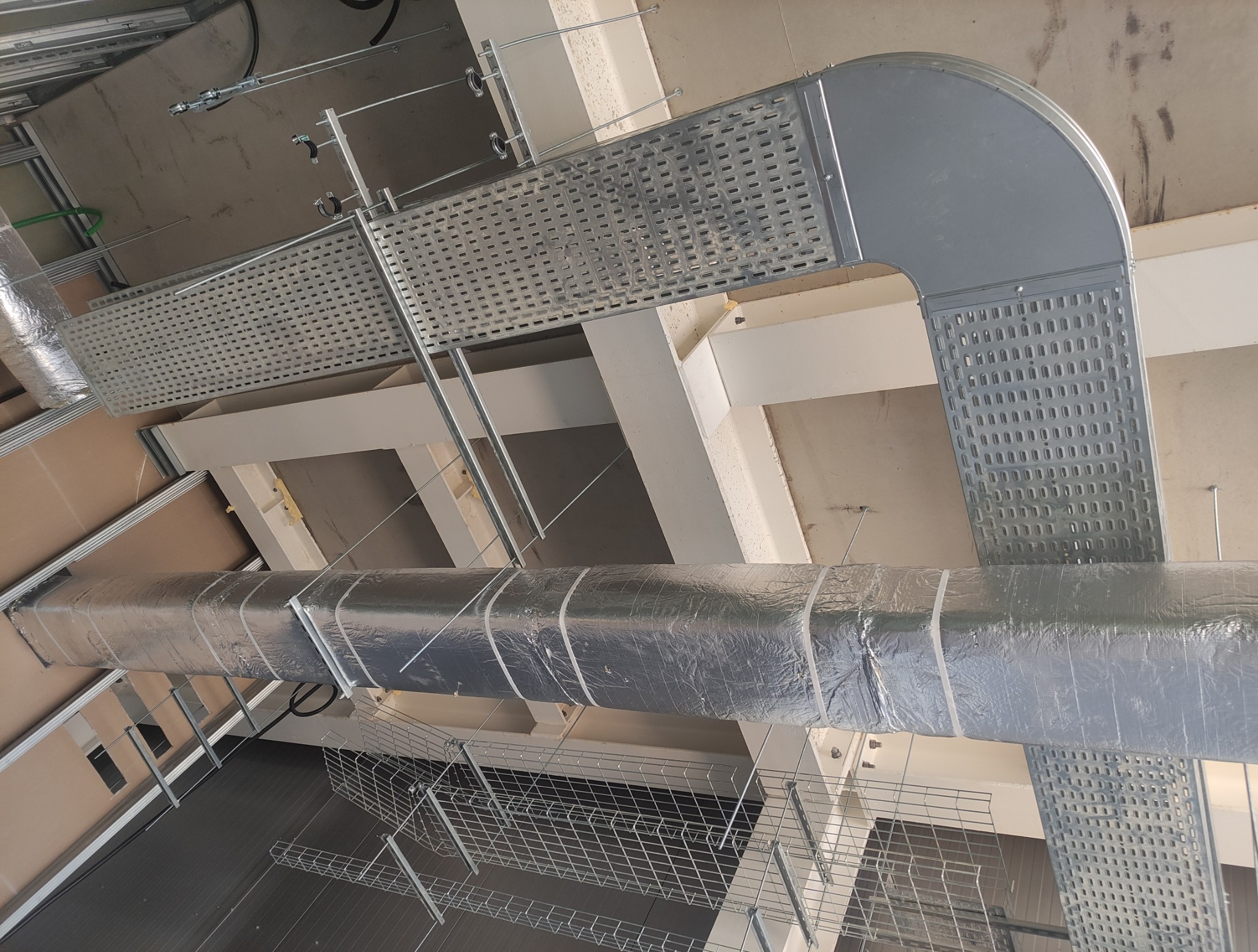 Srebrne koryta kablowe perforowane i izolowany przewód wentylacyjny pod sufitem w budynku. Widok z dołu, instalacje w stanie surowym.