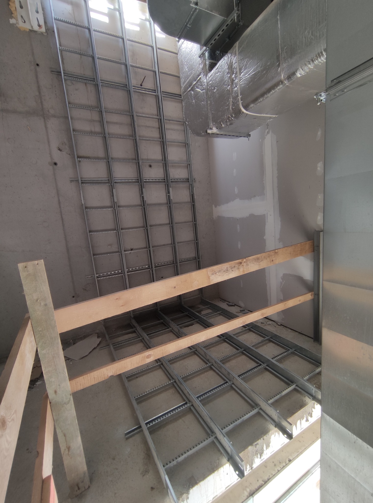 Montaż metalowej konstrukcji pod sufitem, widoczne elementy wentylacji i drewniane podpory. Surowy stan wykończenia wnętrza.