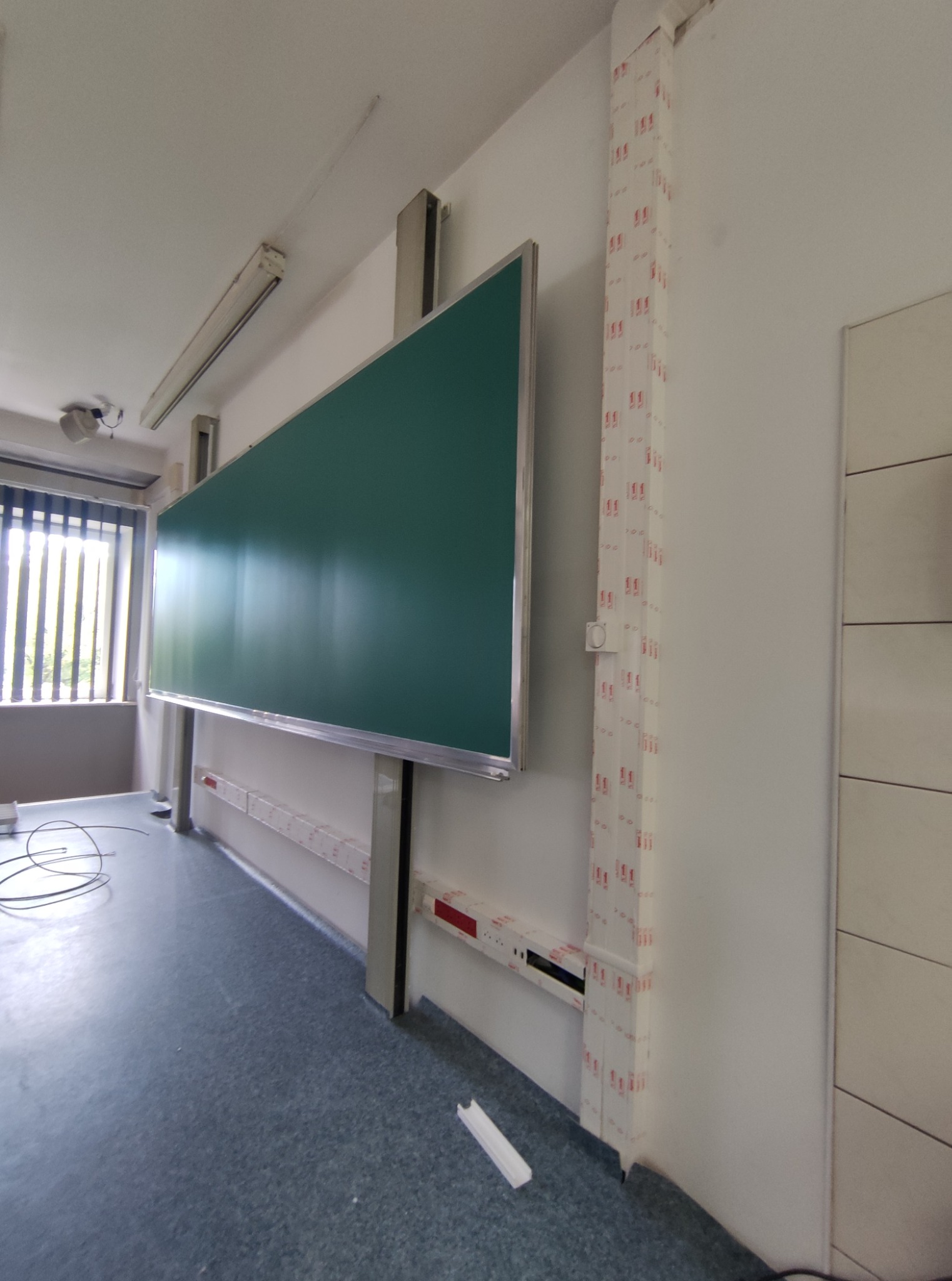 Sala lekcyjna z tablicą i listwami przypodłogowymi. Widoczne kable i elementy instalacji elektrycznej w białych maskownicach na ścianie. Oświetlenie sufitowe.