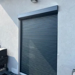 Zamknięta, grafitowa osłona okienna na tle białej ściany budynku, widziana z boku, z delikatnym cieniem i lampą na elewacji.