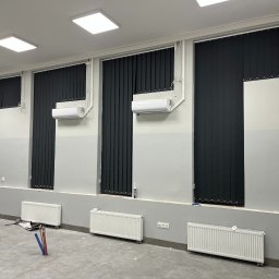 HG Montaż Hubert Głąb - Ciemne, pionowe osłony okienne zamontowane w szarym pomieszczeniu z grzejnikami i klimatyzacją. Widoczne elementy instalacji grzewczej w podłodze.