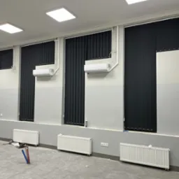 Ciemne, pionowe osłony okienne zamontowane w szarym pomieszczeniu z grzejnikami i klimatyzacją. Widoczne elementy instalacji grzewczej w podłodze.