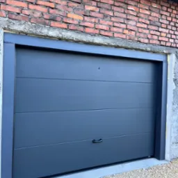 Nowoczesna, szara brama garażowa segmentowa z poziomymi przetłoczeniami, zamontowana w surowym, ceglanym murze. Widoczne detale wykończenia i kamienny podjazd.