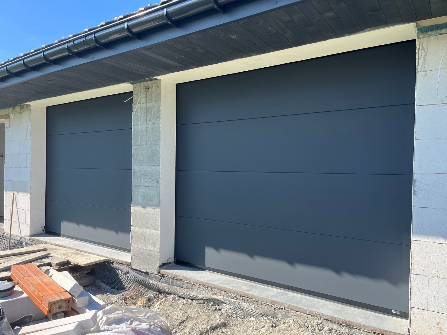 Dwie segmentowe bramy garażowe w kolorze grafitowym, zamontowane w surowym otoczeniu budowy, z widocznymi bloczkami i elementami konstrukcyjnymi.