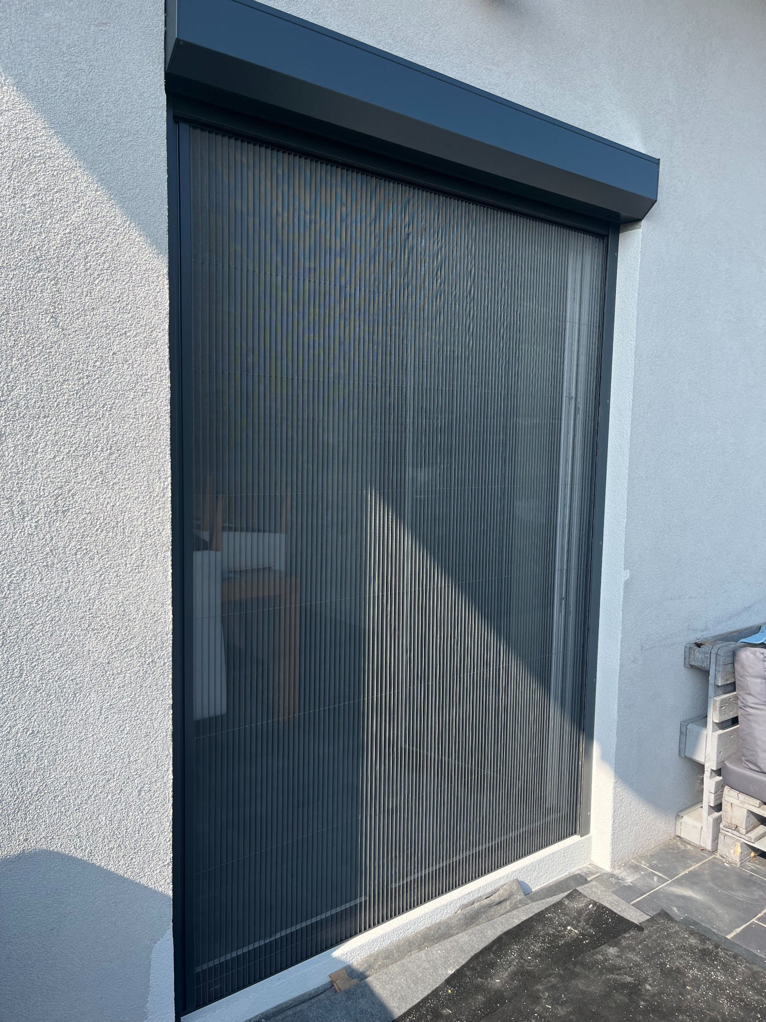 Elegancka, grafitowa moskitiera plisowana, zamontowana na tle białej, teksturowanej ściany zewnętrznej. Widoczne detale konstrukcyjne i subtelne załamania światła.