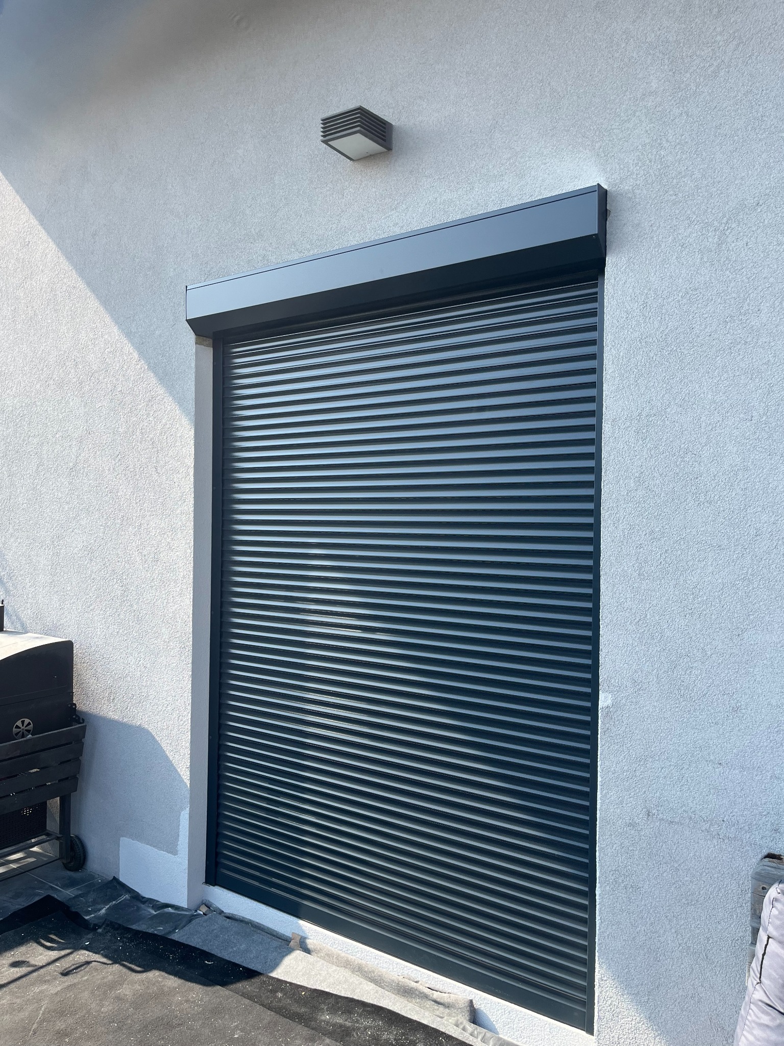 Zamknięta, grafitowa osłona okienna na tle białej ściany budynku, widziana z boku, z delikatnym cieniem i lampą na elewacji.
