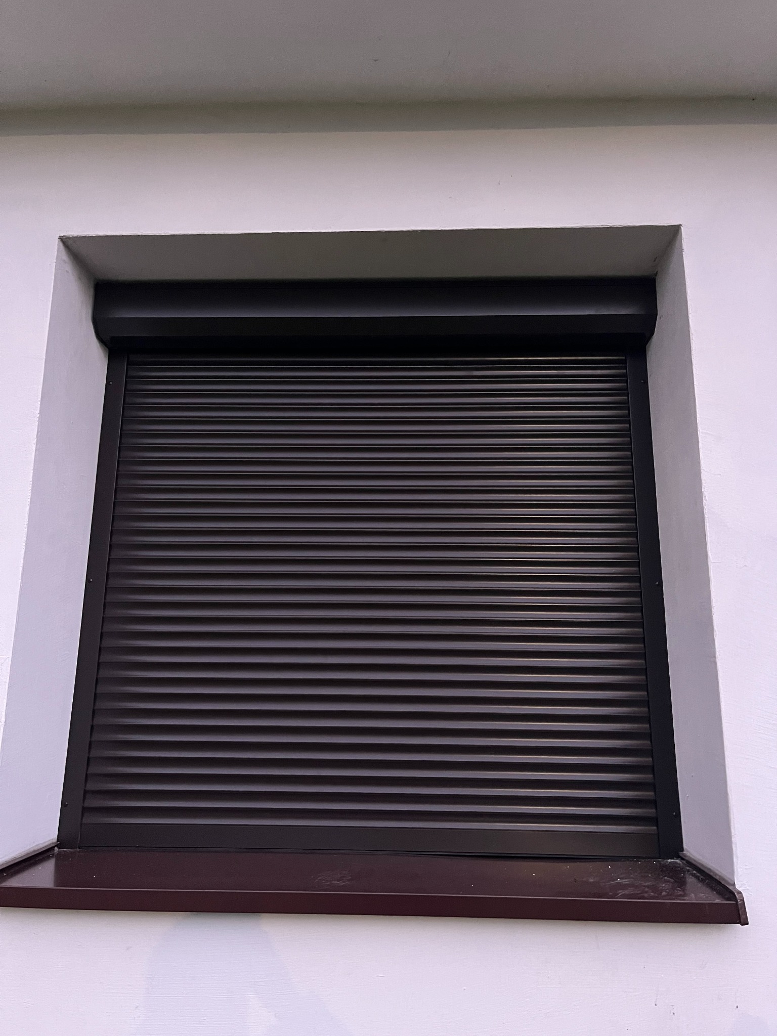 Zasłonięte, brązowe rolety okienne zewnętrzne na tle białej fasady budynku. Widoczny parapet w kolorze zbliżonym do rolet.