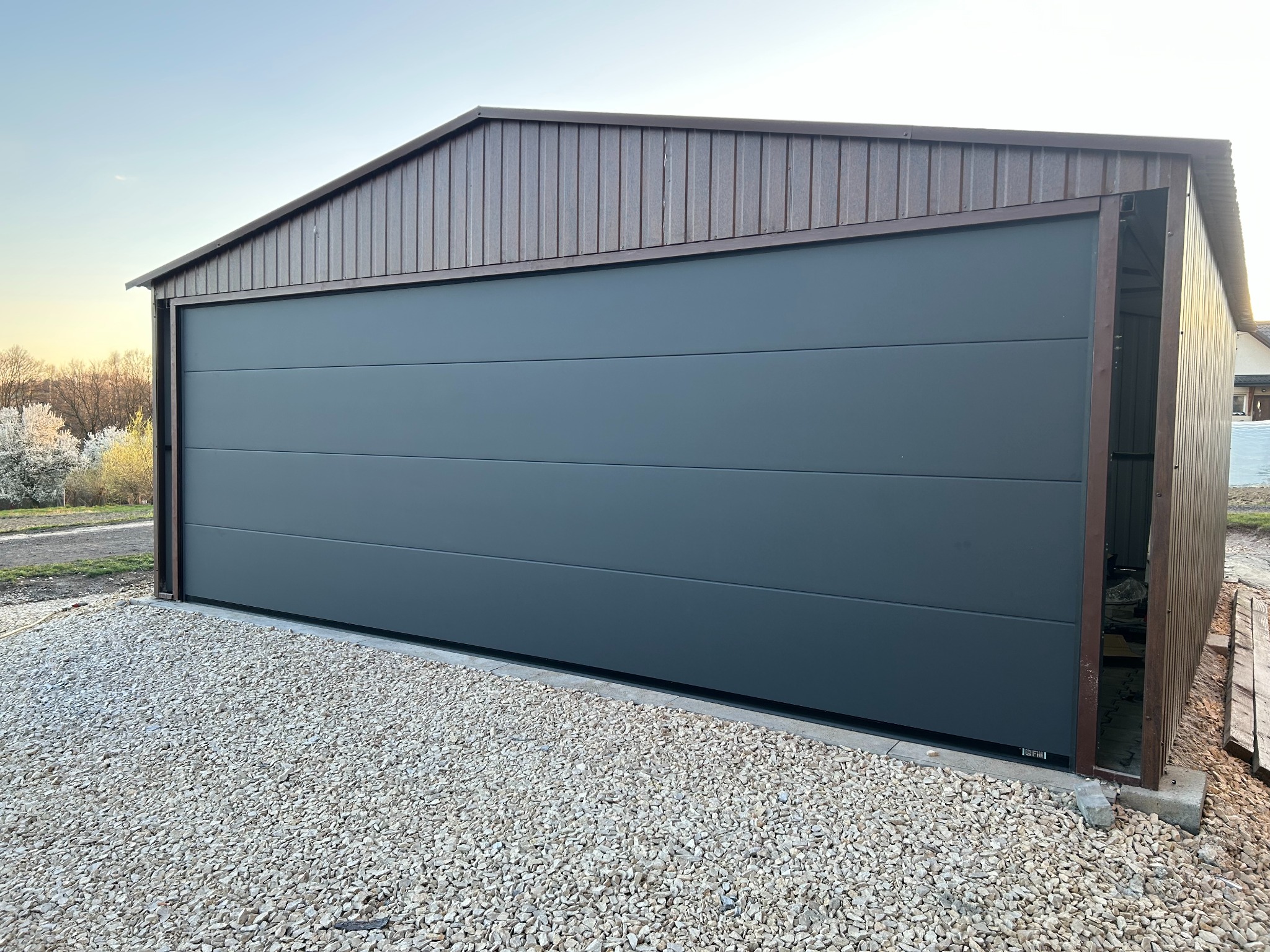 Nowoczesna, segmentowa brama garażowa w kolorze grafitowym, częściowo otwarta, z brązowym obramowaniem i obudową. Podłoże wysypane białym kamieniem.
