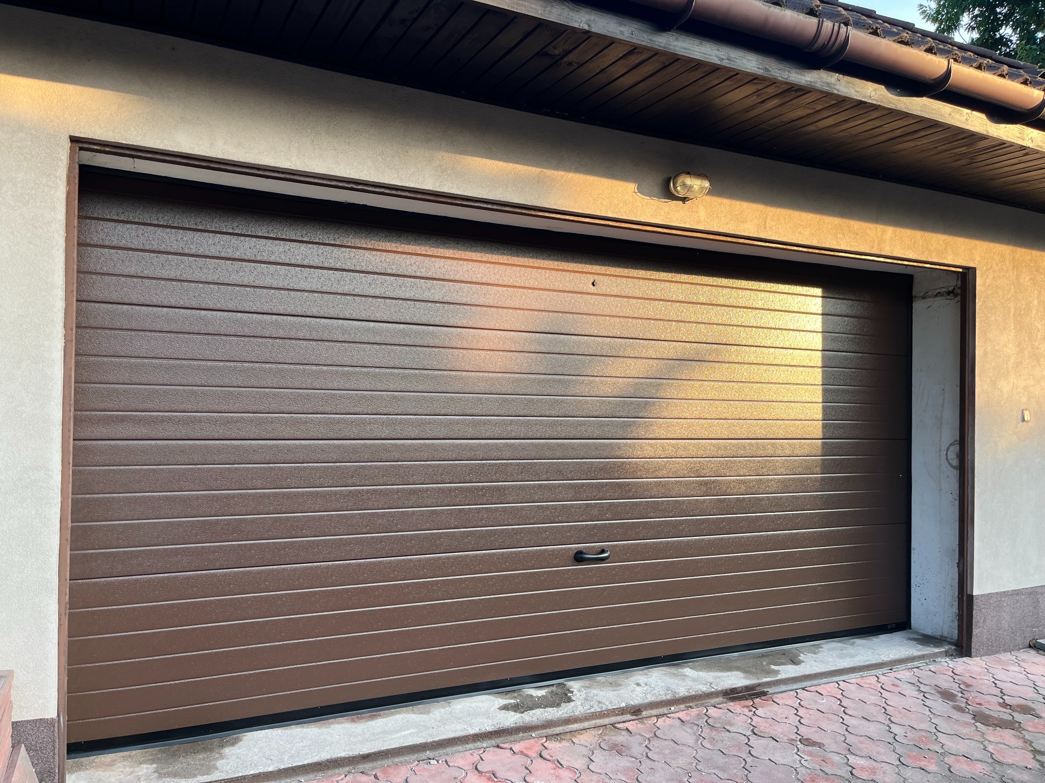 Zamknięta, brązowa brama garażowa segmentowa, oświetlona ciepłym światłem słonecznym, z widoczną lampą zewnętrzną i kostką brukową przed wjazdem.