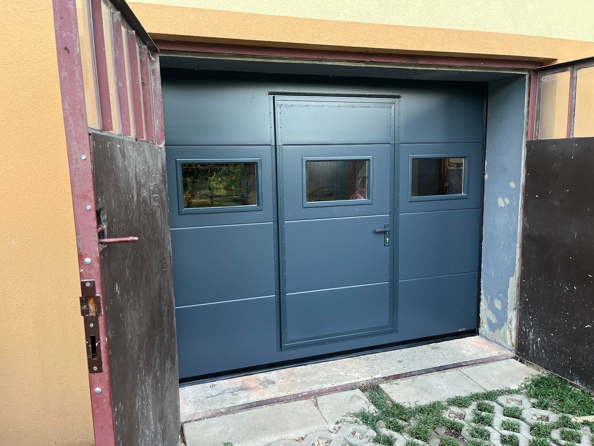 Szara segmentowa brama garażowa z oknami i drzwiami pośrodku, częściowo otwarta, z widoczną fakturą ściany i kostką brukową na podjeździe.