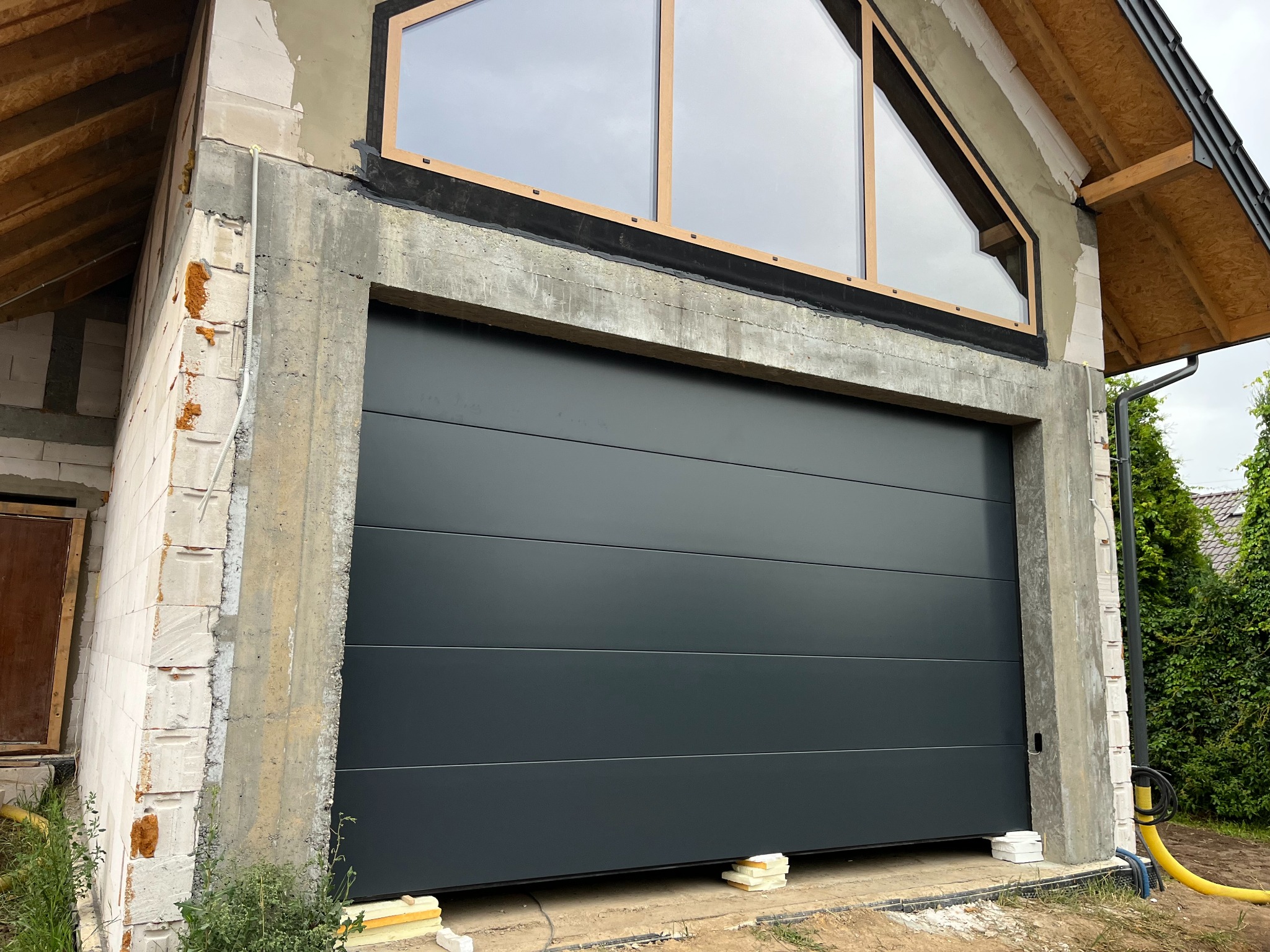 Nowoczesna, segmentowa brama garażowa w kolorze grafitowym, zamontowana w surowym otworze budynku w trakcie budowy, z widocznymi elementami konstrukcyjnymi i oknem trójkątnym.