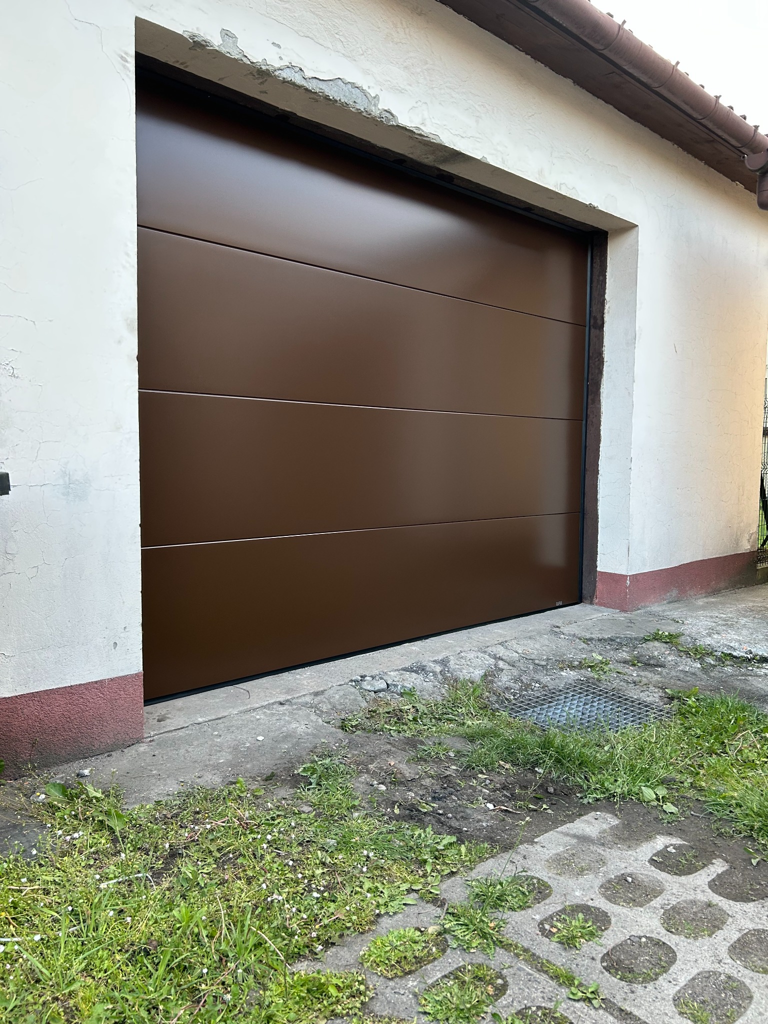 Czekoladowa brama garażowa segmentowa z poziomymi przetłoczeniami, zamontowana w budynku z lekko uszkodzonym tynkiem, widok z zewnątrz.
