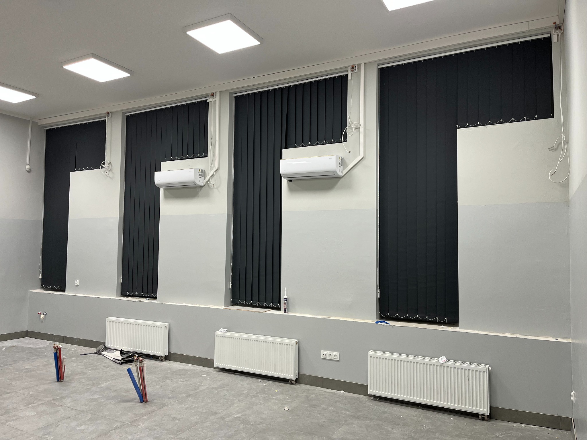 Ciemne, pionowe osłony okienne zamontowane w szarym pomieszczeniu z grzejnikami i klimatyzacją. Widoczne elementy instalacji grzewczej w podłodze.