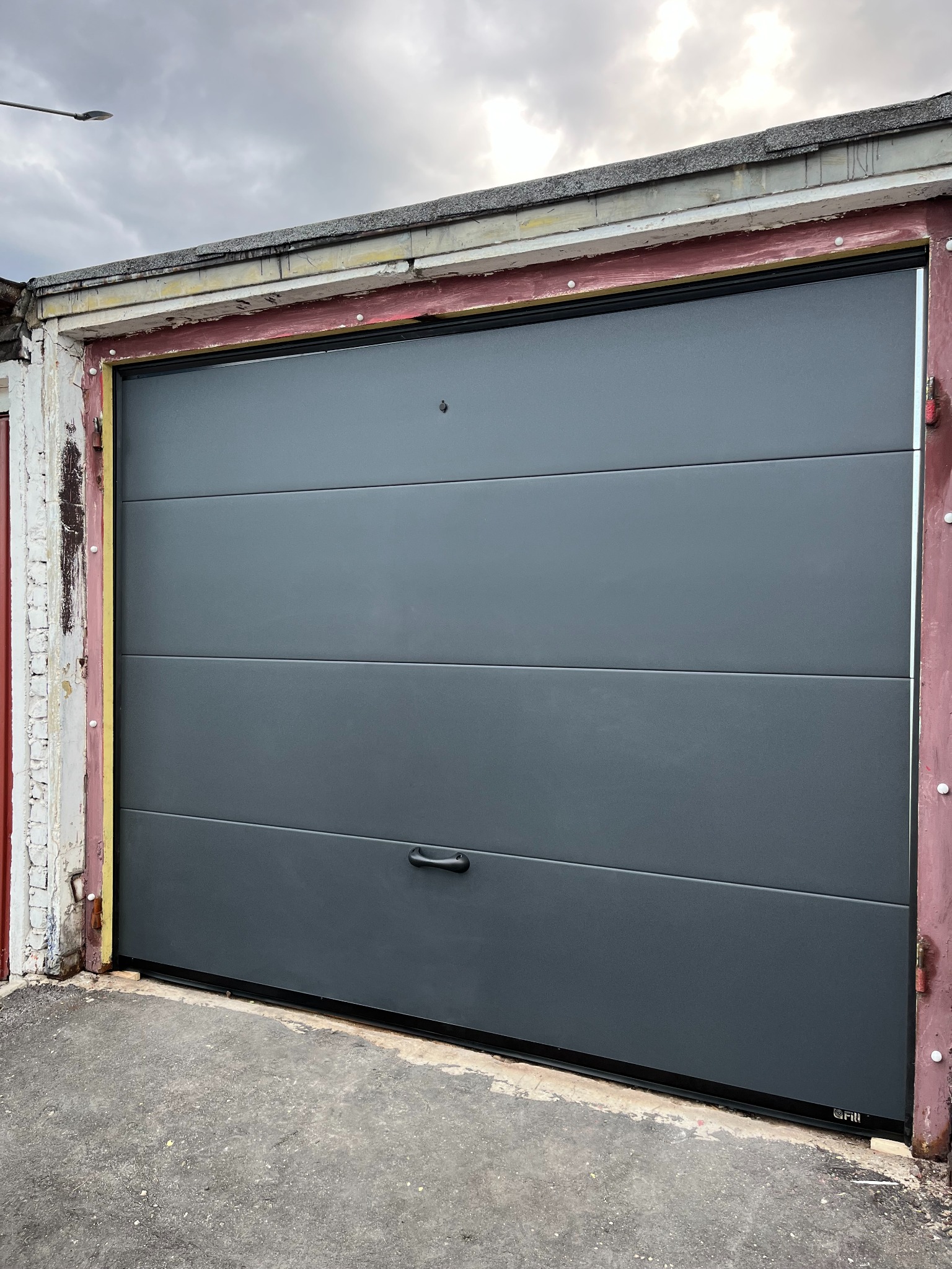 Szara segmentowa brama garażowa z poziomymi panelami, zamontowana w starej, odrapanej ramie. Widoczne elementy mocujące i fragment chodnika.