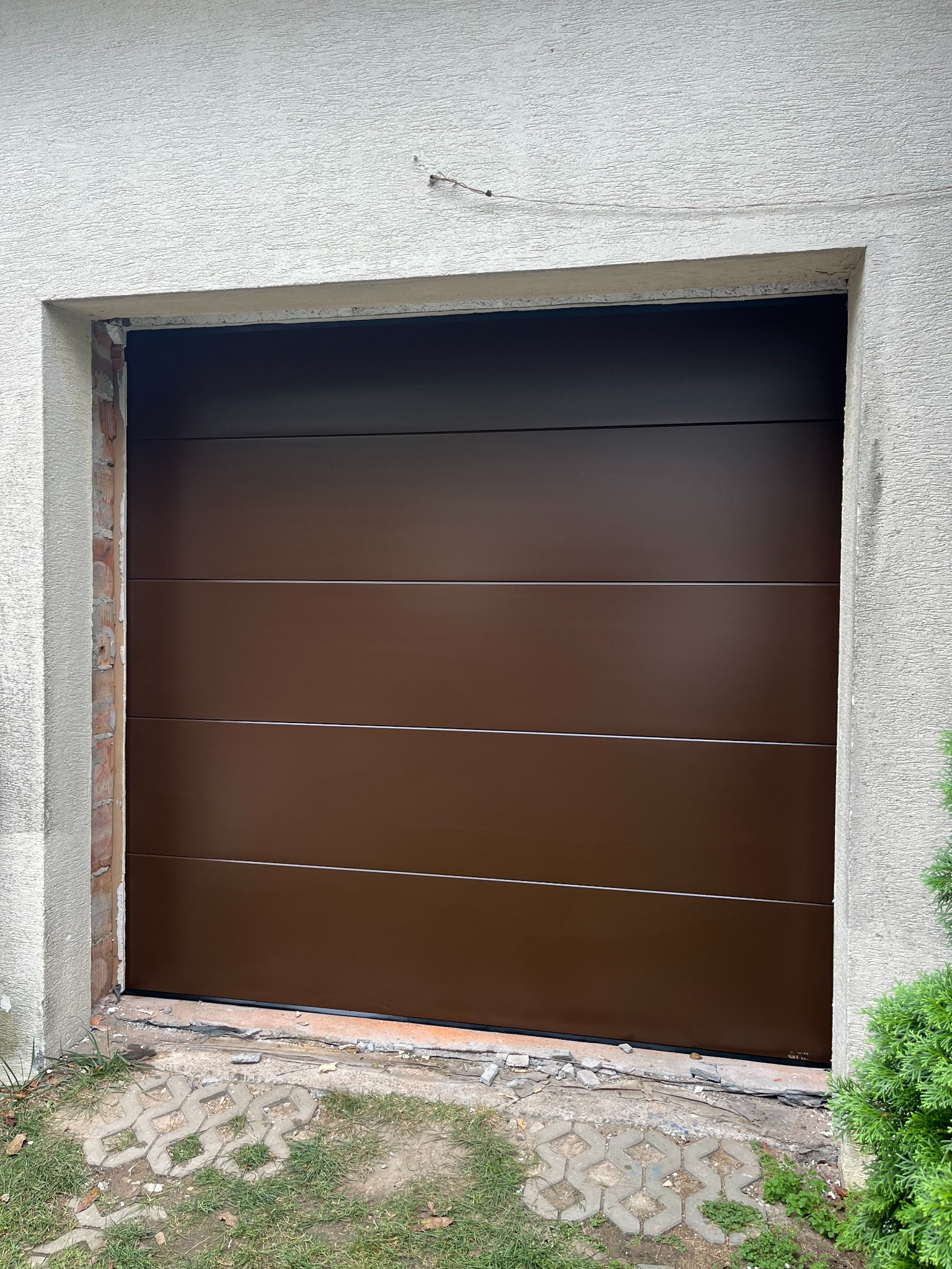 Brązowa segmentowa brama garażowa z poziomymi przetłoczeniami, zamontowana w surowym otworze budynku. Widoczne ślady po montażu i fragment kostki brukowej.