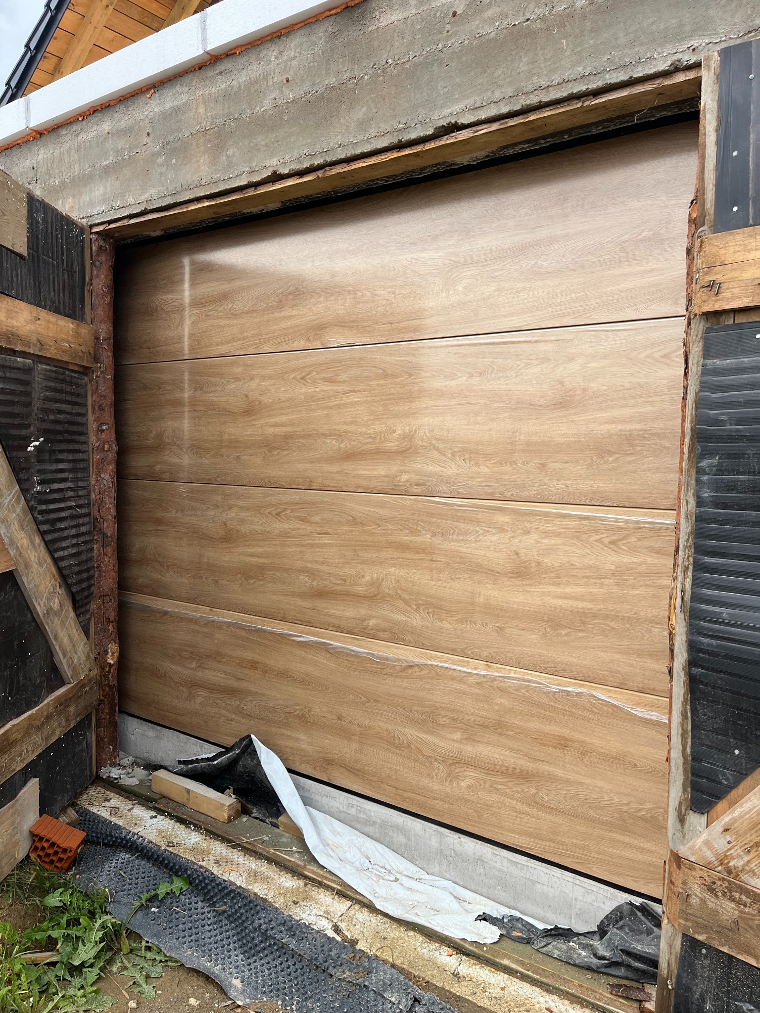 Montaż segmentowej bramy garażowej z wzorem drewna, widoczne elementy konstrukcyjne i zabezpieczenia budowlane. Brama w trakcie instalacji, surowy stan otoczenia.