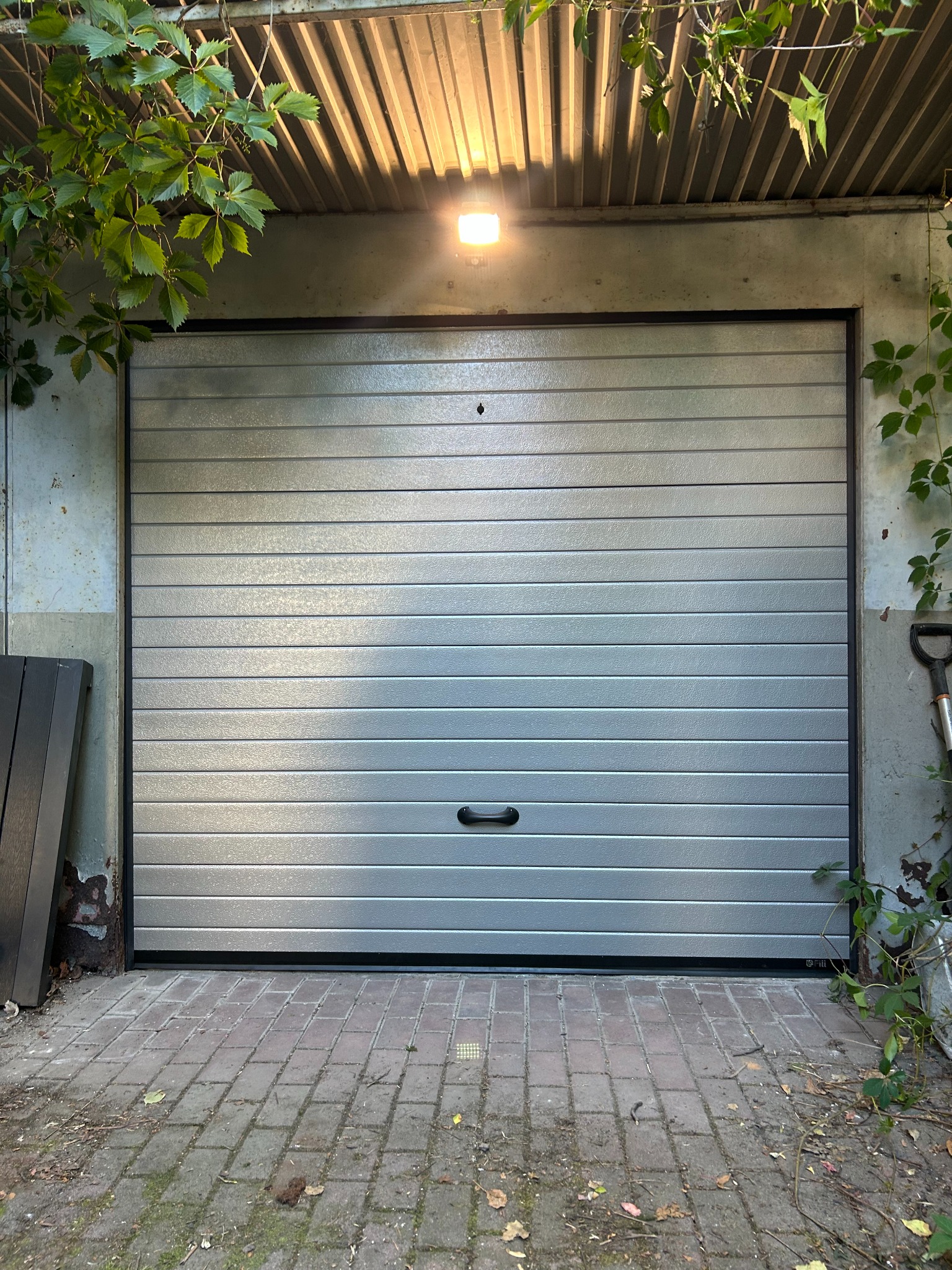 Zamknięta, pozioma, srebrna brama garażowa z czarną rączką na tle ceglanej kostki i zieleni. Nad bramą widoczna lampa oświetlająca wjazd do garażu.