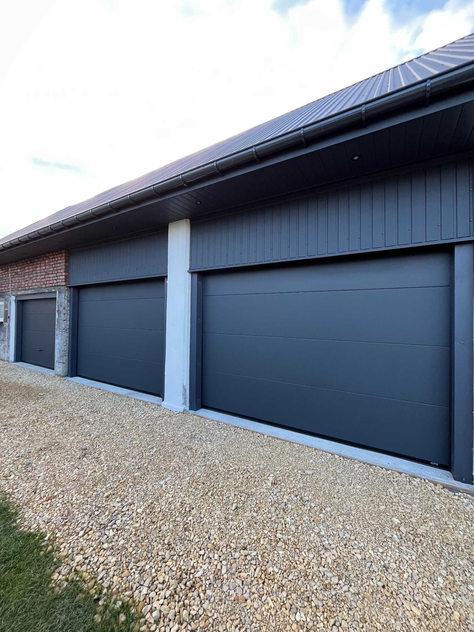 Nowoczesny garaż z trzema segmentowymi bramami w kolorze grafitowym, wykończony kamiennym podłożem i ceglaną ścianą, oświetlony naturalnym światłem.