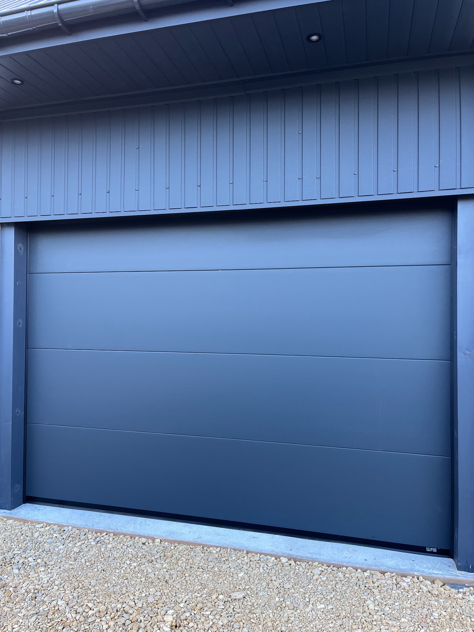 Zamknięta, segmentowa brama garażowa w kolorze antracytowym, z widoczną strukturą paneli i drobnym żwirem przed wjazdem, uchwycona w dziennym świetle.