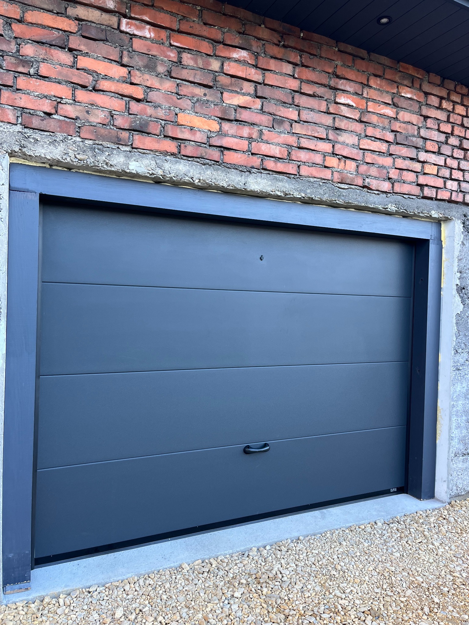 Nowoczesna, szara brama garażowa segmentowa z poziomymi przetłoczeniami, zamontowana w surowym, ceglanym murze. Widoczne detale wykończenia i kamienny podjazd.