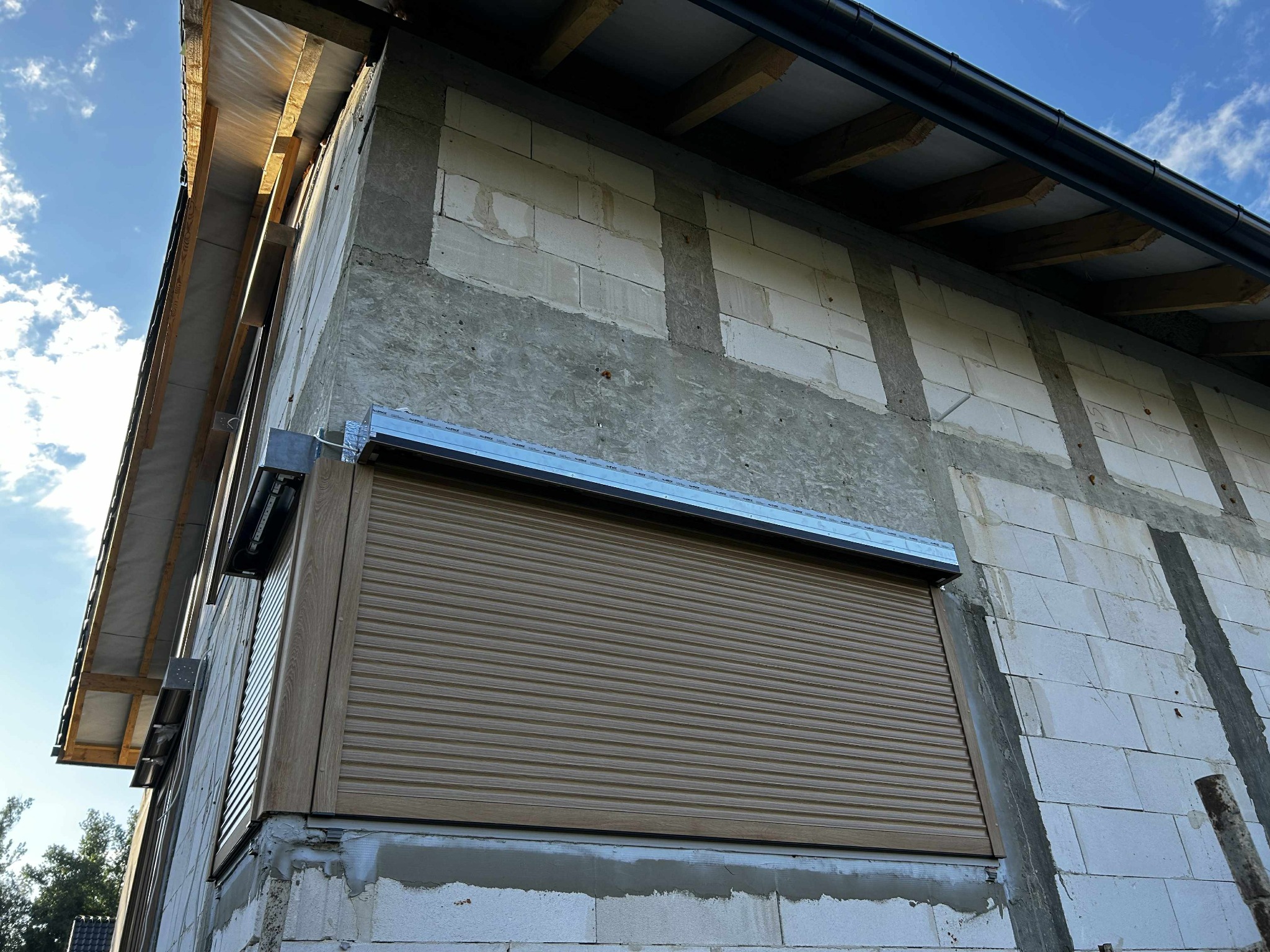 Zamknięta, brązowa roleta na oknie w surowym budynku z białego bloczka, widoczna z dołu pod kątem. Montaż w trakcie budowy.