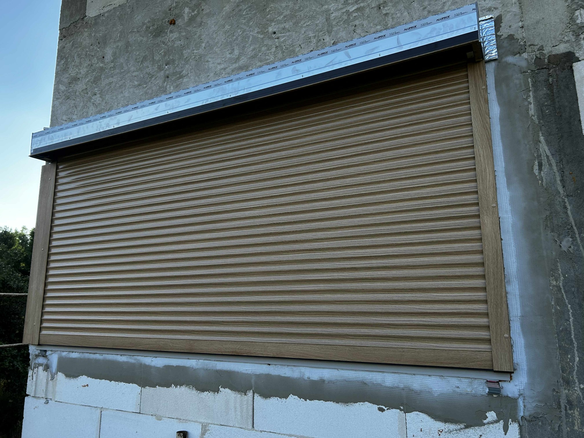 Zamknięta, beżowa osłona okienna z widoczną strukturą drewna, zamontowana na surowej elewacji budynku w trakcie wykończenia.
