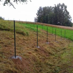 Nazar Rud - Etap montażu ogrodzenia na nierównym terenie: zielone słupki ogrodzeniowe osadzone w betonie, wyznaczające linię przyszłego ogrodzenia na zboczu.