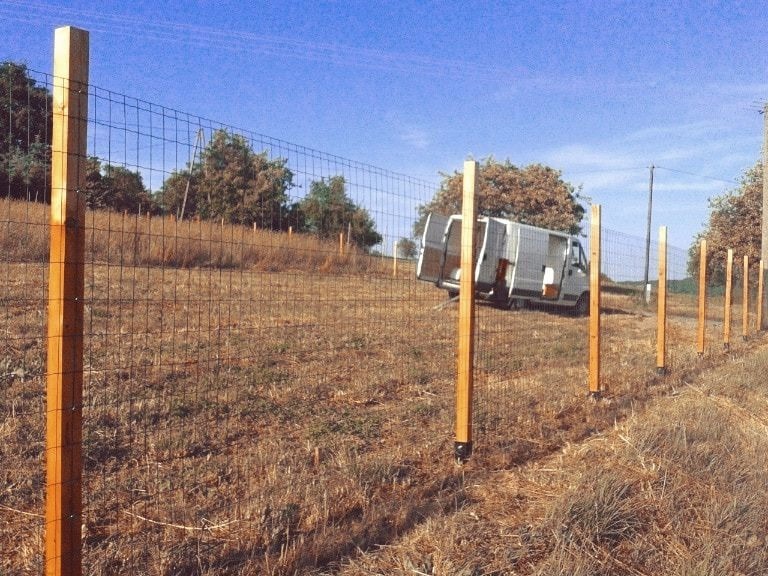 Montaż siatki ogrodzeniowej z drewnianymi słupkami na łące. W tle biały bus z otwartymi drzwiami. Widok na częściowo ukończone ogrodzenie.