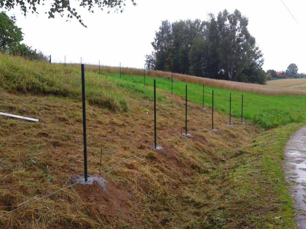 Etap montażu ogrodzenia na nierównym terenie: zielone słupki ogrodzeniowe osadzone w betonie, wyznaczające linię przyszłego ogrodzenia na zboczu.