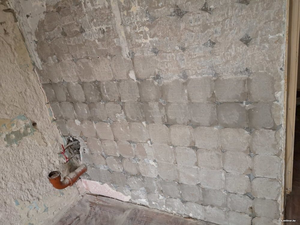 Przygotowana ściana do malowania z widocznymi śladami po usuniętych płytkach i instalacją hydrauliczną. Surowy stan wnętrza przed remontem.