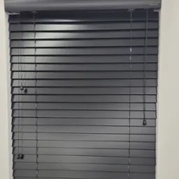 Czarne poziome żaluzje z widocznymi sznurkami i obciążnikami na tle białej ściany, z delikatnym oświetleniem sufitowym.