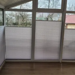 Zasłonięte okna w oranżerii z widokiem na domy za pomocą szarych, plisowanych osłon okiennych.