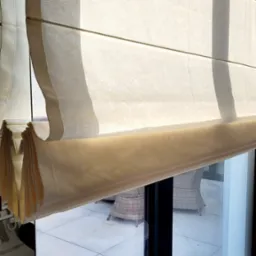 Jasna, kremowa tkanina w systemie fałdowym, tworząca efektowne rolety w oknie z czarną ramą, widok na taras z meblami ogrodowymi.