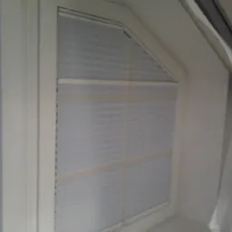 Jasnoszara, dopasowana plisa okienna w nietypowym oknie na poddaszu. Widoczne delikatne poziome fałdy i biała rama okienna. Stonowana kolorystyka.