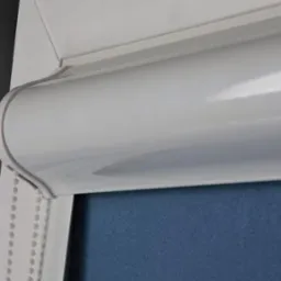 Biała roleta wewnętrzna w kasecie z niebieskim materiałem, widoczna z bliska, z łańcuszkiem sterującym po lewej stronie.