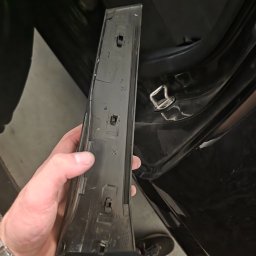 Seweryn kacprzak - Czarny plastikowy element wykończeniowy wnętrza auta, trzymany w dłoni, z widocznymi zaczepami mocującymi. W tle fragment wnętrza pojazdu i podłoga.