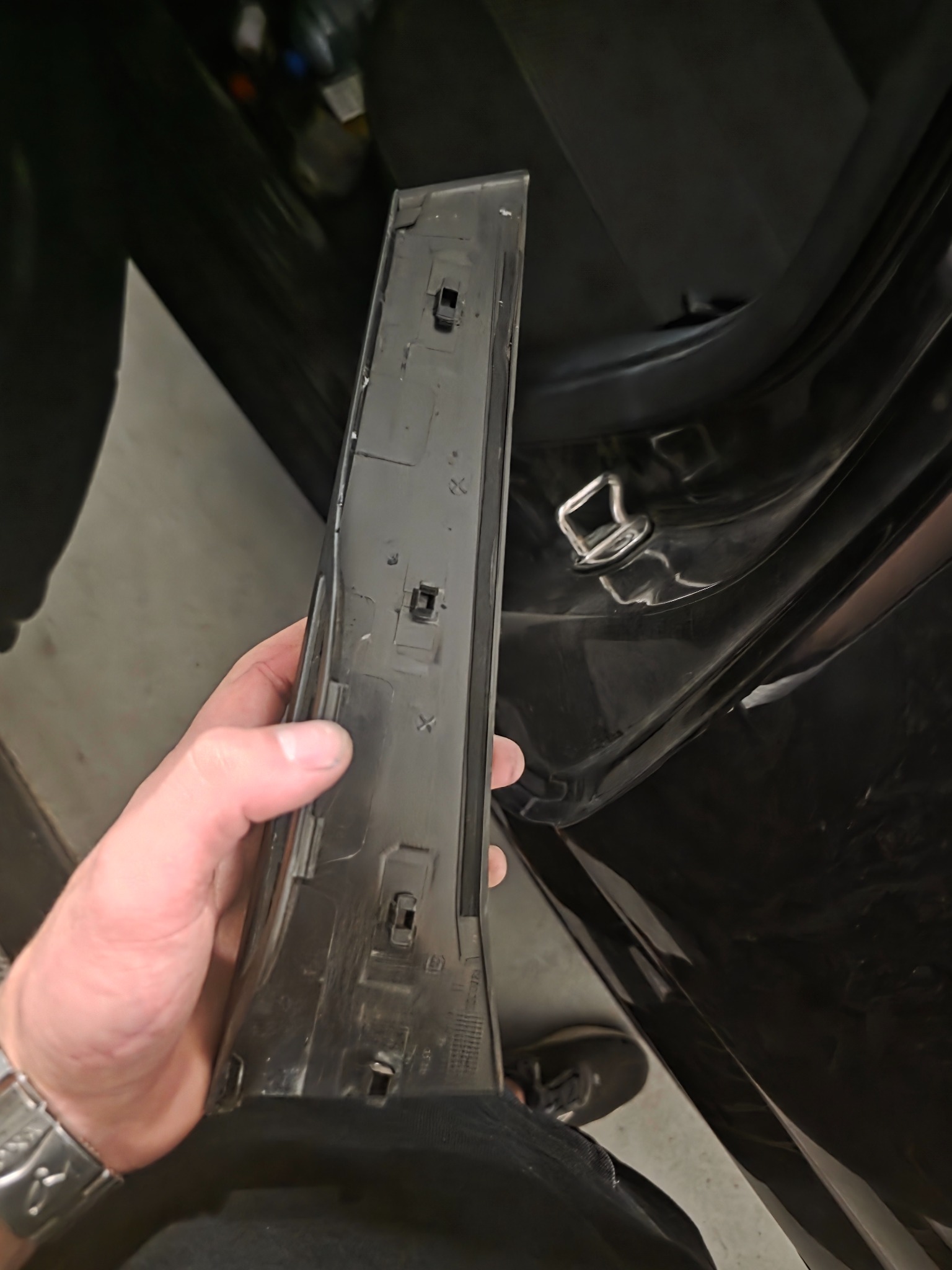 Czarny plastikowy element wykończeniowy wnętrza auta, trzymany w dłoni, z widocznymi zaczepami mocującymi. W tle fragment wnętrza pojazdu i podłoga.