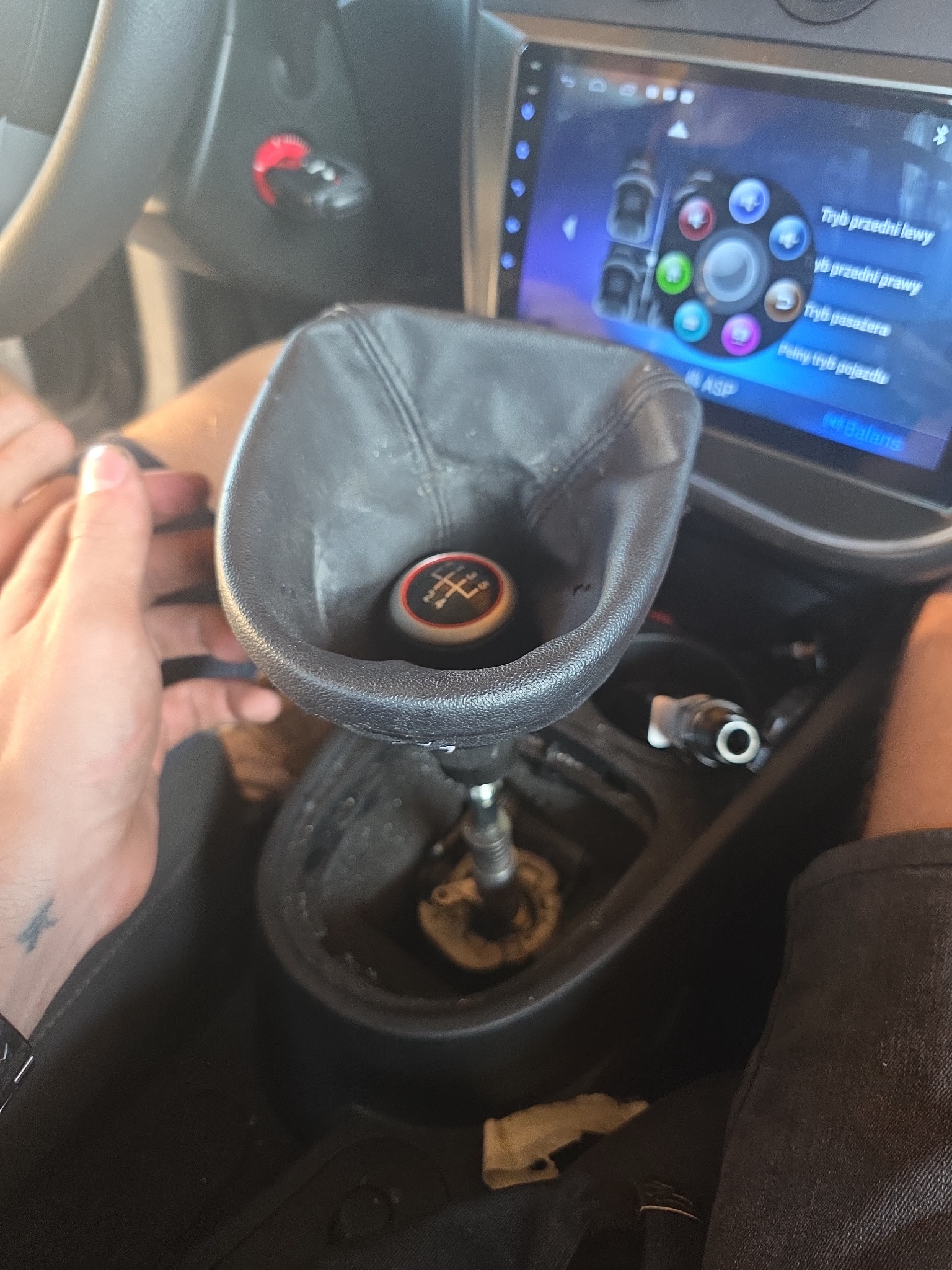 Wnętrze auta: uszkodzony mieszek zmiany biegów z gałką, widoczne elementy deski rozdzielczej i panel sterowania. Detal z naprawy lub wymiany.