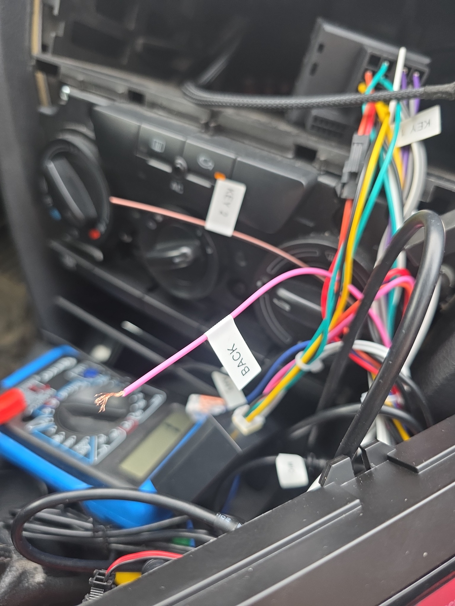 Podłączenie przewodów w samochodzie, widoczne kolorowe kable z etykietami, miernik elektryczny. Prace instalacyjne w desce rozdzielczej pojazdu.