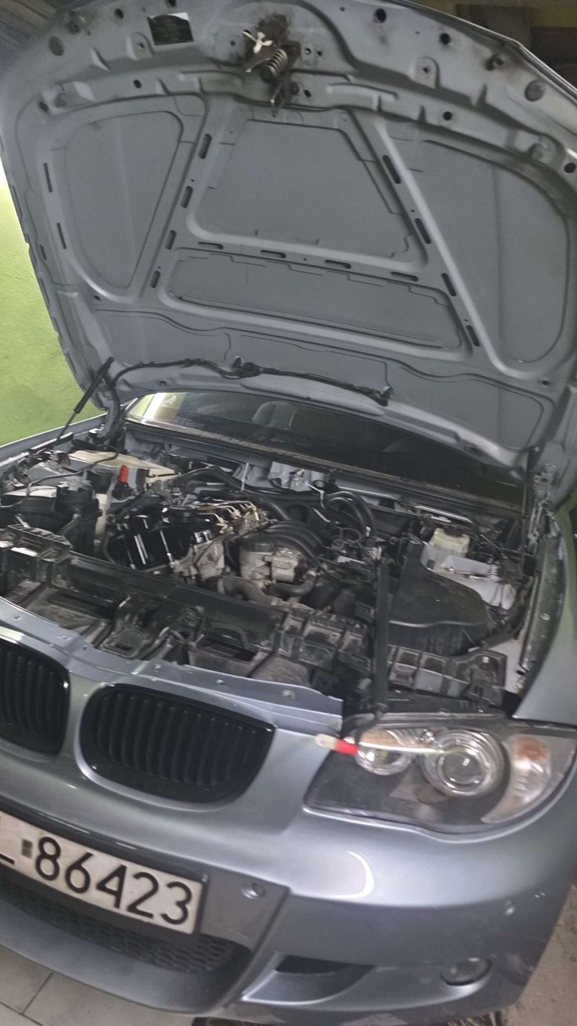 Otwarta maska srebrnego samochodu z widocznym silnikiem w trakcie naprawy. Widoczne przewody i elementy mechaniczne. Tablica rejestracyjna z przodu pojazdu.