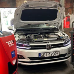 Biały samochód z otwartą maską w warsztacie, widoczna maszyna do klimatyzacji TEXA 705R R134a i wózek narzędziowy ROCKS. Tablica rejestracyjna SBI 1909F.